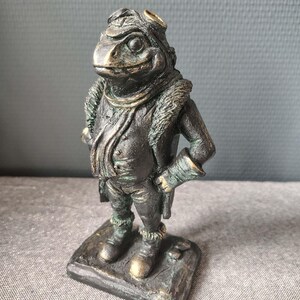 May include: A bronze-toned figurine of an anthropomorphic toad wearing an aviator cap with goggles, a collared jacket with fur-like trim, and boots. The toad stands on a rectangular base with a weathered, metallic finish and textured details.