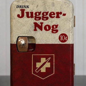 Op de afbeelding: Een rechthoekige metalen automaat-replica met een verweerde crème en donkerrode afwerking. De tekst luidt DRINK Jugger-Nog en 10 cent. Een gouden kruis-embleem met een diagonale lijn is gecentreerd op het onderste rode gedeelte met een uitsparing.