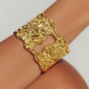May include: A gold cuff bracelet with a textured, organic design. The bracelet features a large, open centre and a wavy, sculpted surface. The jewellery is worn on a wrist, showcasing its intricate details and metallic sheen.