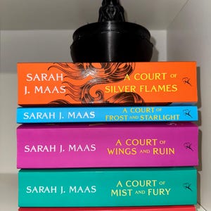 Può includere: Una pila di cinque libri di Sarah J. Maas, ognuno con una copertina di colore diverso. I titoli includono "A Court of Silver Flames", "A Court of Frost and Starlight" e "A Court of Wings and Ruin". Un oggetto decorativo nero è in cima.