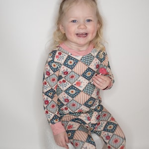 May include: A child's two-piece pajama set with a floral patchwork design. The long-sleeved top and pants feature a pattern of flowers, diamonds, and stripes in shades of pink, blue, and beige. The pajamas have a pink neckline and cuffs.