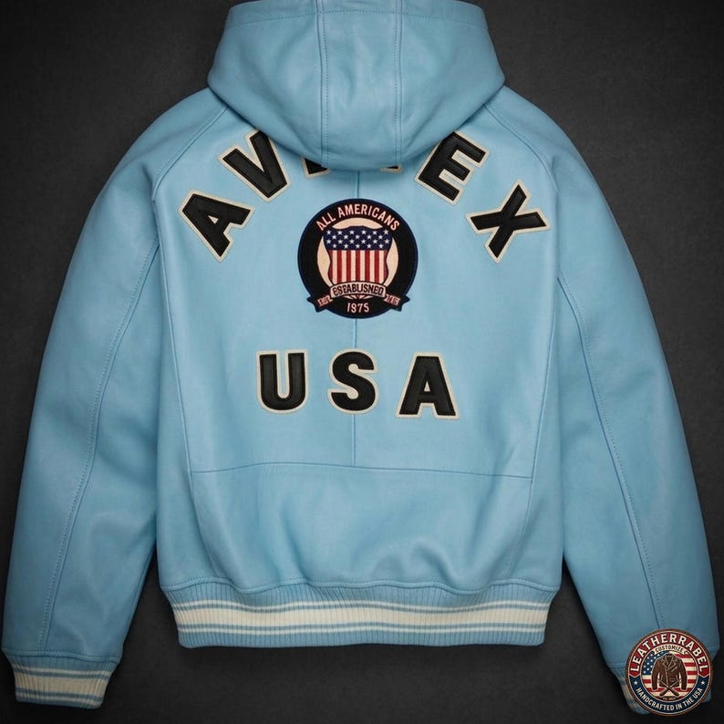 Pu&ograve; includere: Giacca in pelle blu chiaro con cappuccio. La schiena presenta la scritta "AVIREX" in lettere nere, con "USA" sotto una toppa circolare con un disegno della bandiera americana. La giacca ha un bordo a righe bianche e blu in vita.