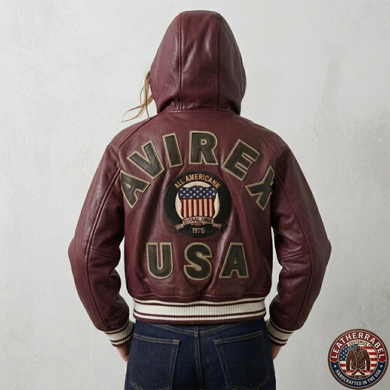 Puede incluir: Una chaqueta bomber de cuero color burdeos con capucha que presenta el texto AVIREX USA en letras grandes y oscuras en la espalda. Un parche circular en el centro muestra un gr&aacute;fico de la bandera estadounidense con el texto ALL AMERICAN ESTAAL ANNE 1975. La chaqueta tiene pu&ntilde;os y dobladillo acanalados con rayas blancas.