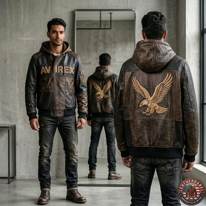 May include: A brown leather hooded jacket with the text "AVIREX" on the chest and an eagle emblem on the back. The jacket has a bomber style with black ribbed cuffs and hem. The model is wearing dark wash jeans and brown boots.