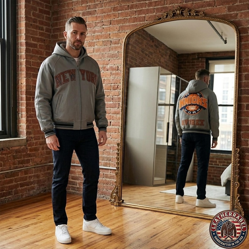 Puede incluir: Una chaqueta gris con capucha y cremallera con el texto NEW YORK en rojo oscuro en el pecho. La parte posterior presenta un gran gr&aacute;fico circular naranja y azul con el texto KNICKS y NEW YORK, junto con la palabra AVOREX debajo. Vaqueros azul oscuro y zapatillas blancas.