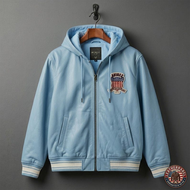 Pu&ograve; includere: Giacca in pelle azzurra con cappuccio e chiusura a zip. La giacca presenta una toppa sul petto con la scritta "AVIREX" e un emblema. I polsini e l'orlo hanno strisce bianche.