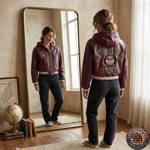 Puede incluir: Una chaqueta bomber de cuero color burdeos con capucha, cremallera frontal, pu&ntilde;os y dobladillo acanalados con rayas blancas. La espalda muestra el texto AVIREX USA en letras grandes rodeando un parche circular con una bandera estadounidense.