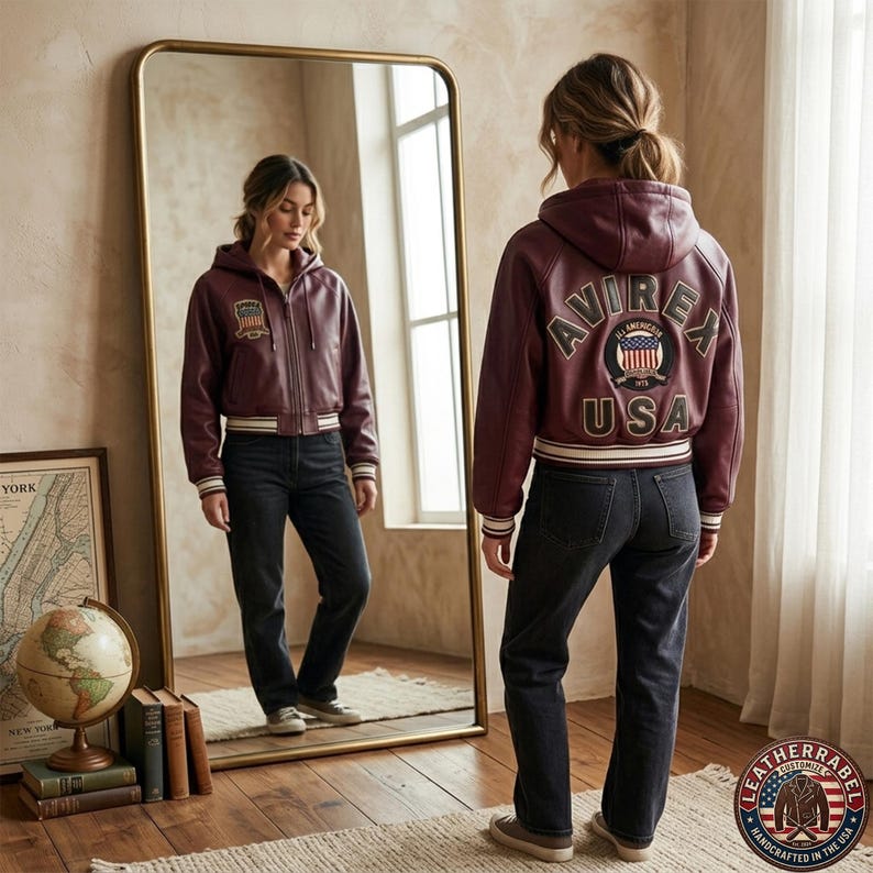 Puede incluir: Una chaqueta bomber de cuero color burdeos con capucha, cremallera frontal, pu&ntilde;os y dobladillo acanalados con rayas blancas. La espalda muestra el texto AVIREX USA en letras grandes rodeando un parche circular con una bandera estadounidense.