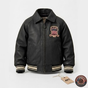 Puede incluir: Chaqueta de cuero negra con cierre de cremallera, cuello y parche con dise&ntilde;o de bandera americana. La chaqueta tiene pu&ntilde;os y dobladillo a rayas. Una etiqueta con el texto "AVI" cuelga.