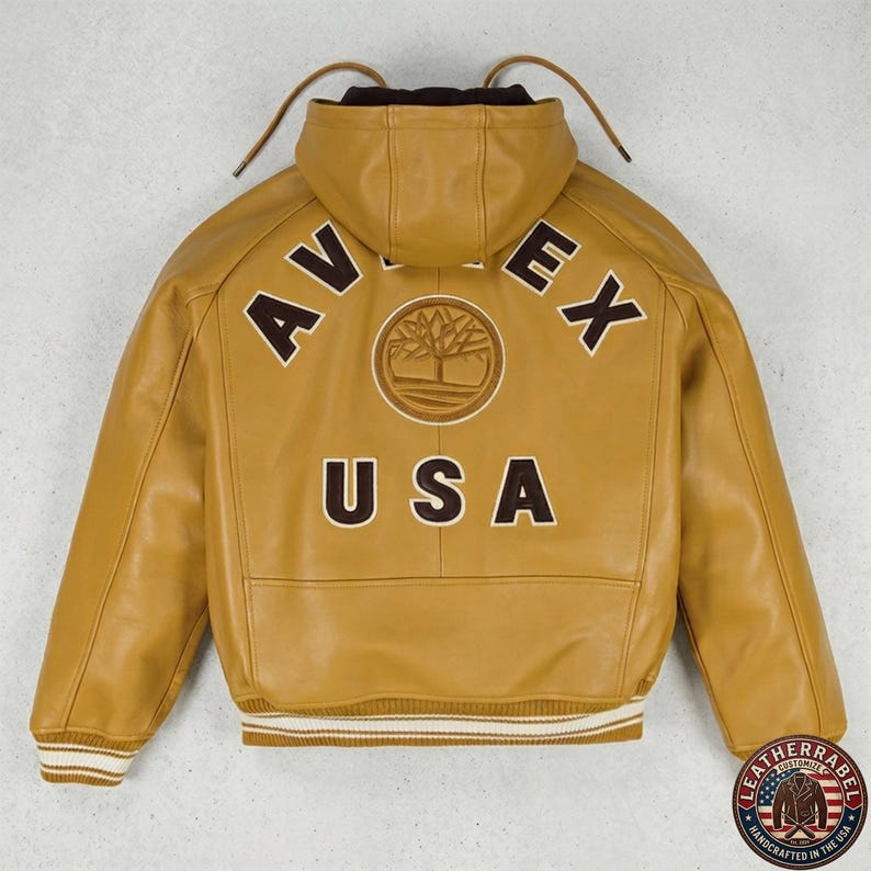 May include: Mustard yellow leather jacket with a hood. The back features the text "AVEX USA" in brown block letters, with a circular emblem. The jacket has a ribbed hem and cuffs with white stripes. A "LEATHERRABEL" logo is visible.