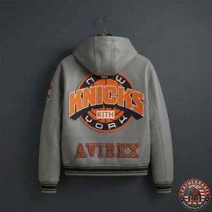 Puede incluir: Una chaqueta de cuero gris con capucha que presenta un gran parche circular en la espalda con el texto New York Knicks y Kith en naranja y azul marino. La palabra Avirex est&aacute; bordada en naranja debajo del parche. Tiene pu&ntilde;os y dobladillo acanalados.