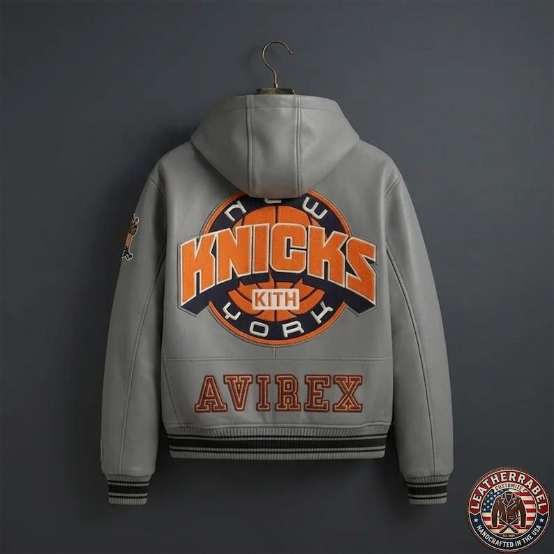 Puede incluir: Una chaqueta de cuero gris con capucha que presenta un gran parche circular en la espalda con el texto New York Knicks y Kith en naranja y azul marino. La palabra Avirex est&aacute; bordada en naranja debajo del parche. Tiene pu&ntilde;os y dobladillo acanalados.