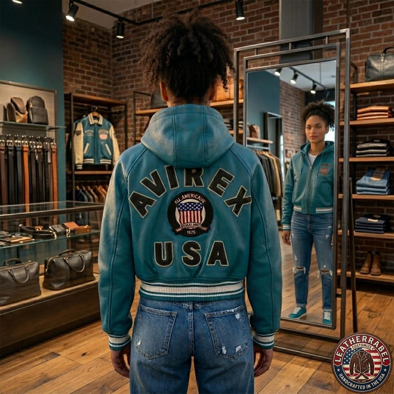 K&ouml;nnte beinhalten: Eine t&uuml;rkisfarbene Leder-Kapuzenjacke im College-Stil mit dem Schriftzug AVIREX USA und einem runden US-Flaggen-Aufn&auml;her auf dem R&uuml;cken. Die Jacke hat gerippte B&uuml;ndchen und einen Saum mit wei&szlig;en und dunklen Streifen und wird zu blauen Jeans getragen.