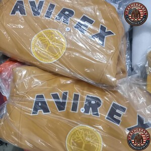 May include: Two tan leather jackets are visible, each wrapped in clear plastic. The jackets feature the word "AVIREX" in large, black block letters across the chest. A circular patch with a tree design is also visible. The jackets are from "LEATHERRABEL".