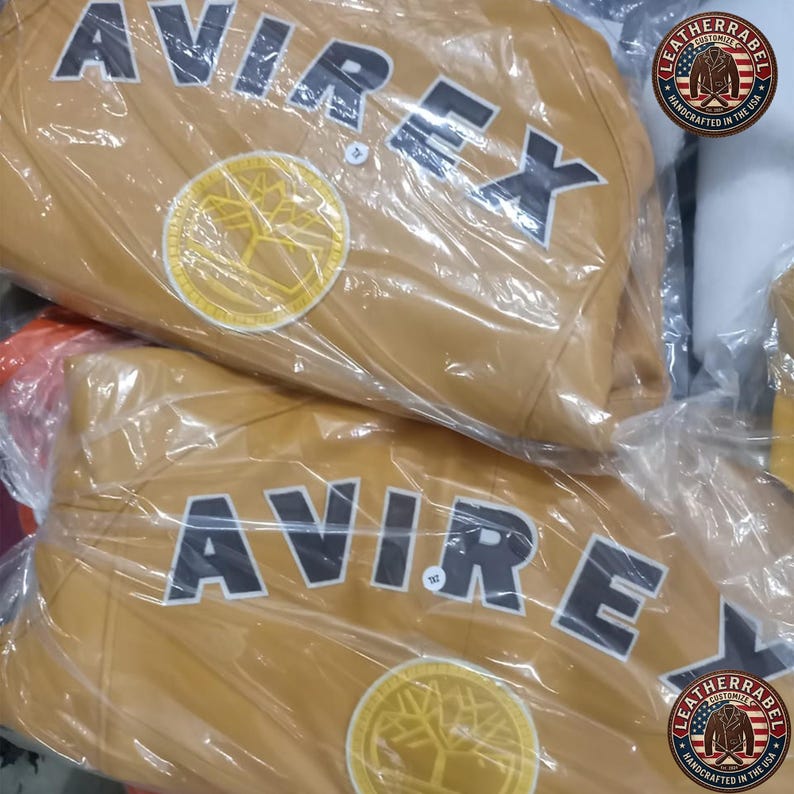 May include: Two tan leather jackets are visible, each wrapped in clear plastic. The jackets feature the word "AVIREX" in large, black block letters across the chest. A circular patch with a tree design is also visible. The jackets are from "LEATHERRABEL".