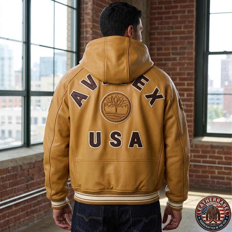May include: Mustard-colored leather jacket with a hood. The back features the word "AVEX" and "USA" in brown lettering, along with a tree logo. The jacket has a ribbed hem and cuffs with white stripes. The Leather Label logo is visible.