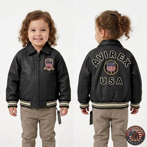 Puede incluir: Chaqueta de cuero negra con el logo "AVIREX USA" bordado en la espalda y un logotipo m&aacute;s peque&ntilde;o en el pecho. La chaqueta tiene cuello, cierre de cremallera y pu&ntilde;os y dobladillo a rayas. El ni&ntilde;o lleva pantalones color caqui.