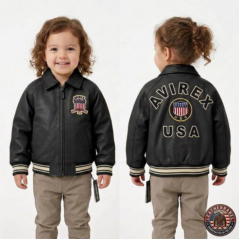 Puede incluir: Chaqueta de cuero negra con el logo "AVIREX USA" bordado en la espalda y un logotipo m&aacute;s peque&ntilde;o en el pecho. La chaqueta tiene cuello, cierre de cremallera y pu&ntilde;os y dobladillo a rayas. El ni&ntilde;o lleva pantalones color caqui.