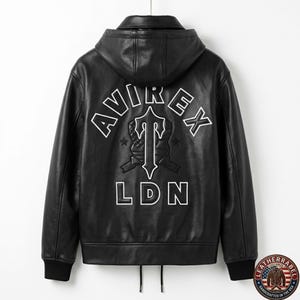 May include: Black leather jacket with a hood. The back features white embroidered text that reads "AVIREX" above a stylized emblem and "LDN" below. The jacket has a zipper closure and black ribbed cuffs and hem.