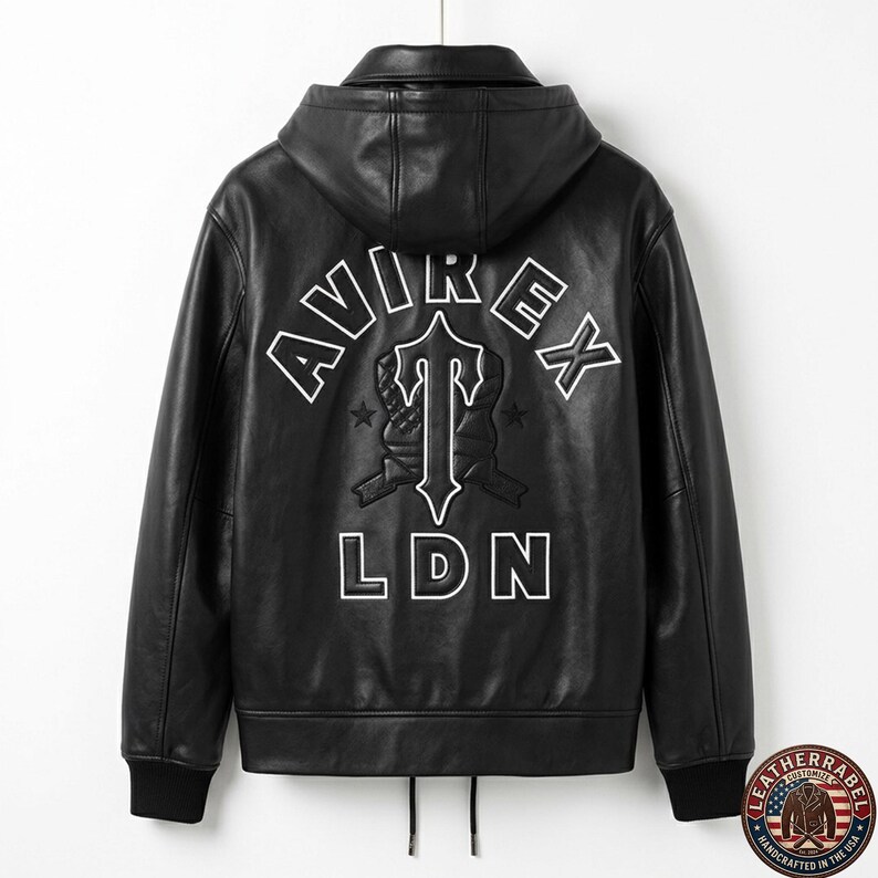 May include: Black leather jacket with a hood. The back features white embroidered text that reads "AVIREX" above a stylized emblem and "LDN" below. The jacket has a zipper closure and black ribbed cuffs and hem.
