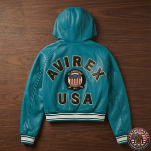 K&ouml;nnte beinhalten: Eine t&uuml;rkisfarbene Lederjacke mit Kapuze und dem Schriftzug AVIREX USA in schwarzen und wei&szlig;en Buchstaben auf der R&uuml;ckseite. Ein runder Aufn&auml;her in der Mitte zeigt eine US-Flagge mit dem Text ALL AMERICANS und 1975. Saum und &Auml;rmelb&uuml;ndchen sind gerippt.