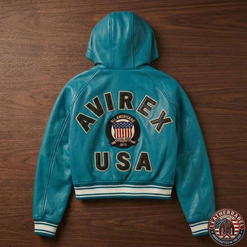 K&ouml;nnte beinhalten: Eine t&uuml;rkisfarbene Lederjacke mit Kapuze und dem Schriftzug AVIREX USA in schwarzen und wei&szlig;en Buchstaben auf der R&uuml;ckseite. Ein runder Aufn&auml;her in der Mitte zeigt eine US-Flagge mit dem Text ALL AMERICANS und 1975. Saum und &Auml;rmelb&uuml;ndchen sind gerippt.