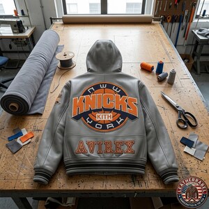 Puede incluir: Una chaqueta universitaria de cuero gris con capucha, que presenta un gran logotipo circular naranja y azul marino de Knicks Kith New York en la espalda. La palabra AVIREX est&aacute; bordada en naranja debajo del logo. Tiene pu&ntilde;os y dobladillo acanalados.