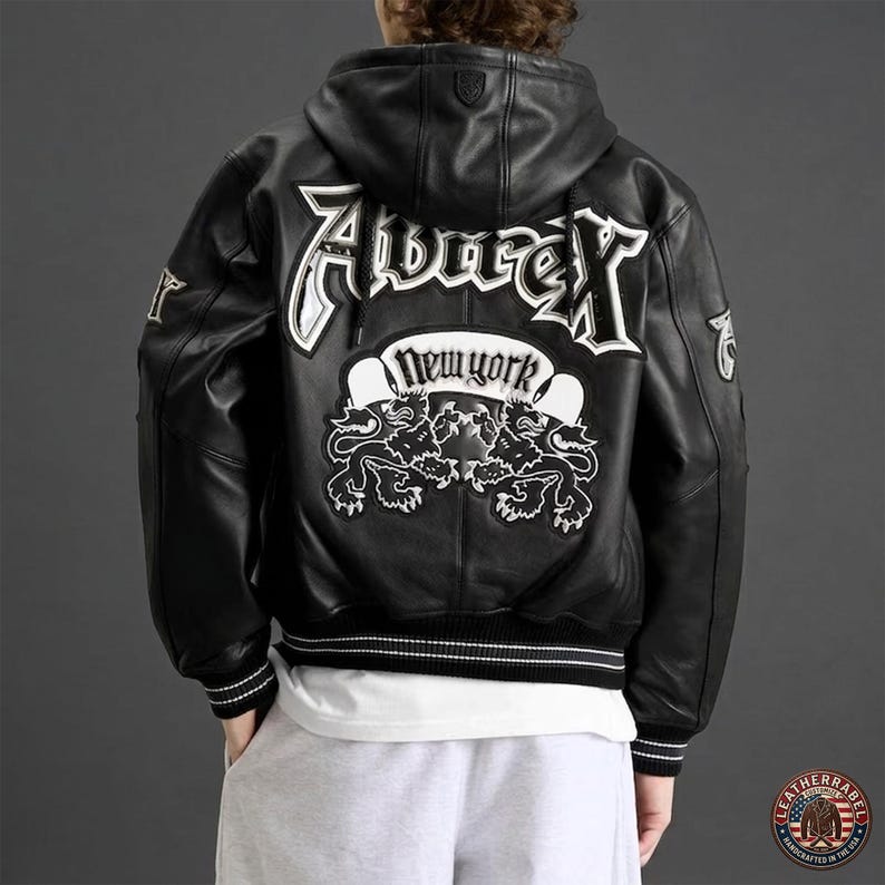 May include: A black leather hooded jacket featuring white embroidered text that reads Avirex and New York, accompanied by two lion graphics on the back. The jacket includes ribbed cuffs and a hem with white stripes, and a small logo on the hood.