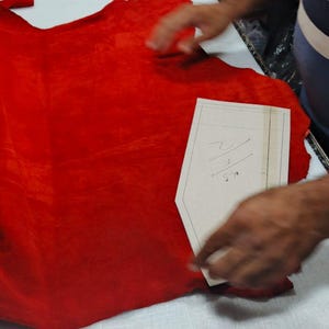 Puede incluir: Una pieza grande de cuero rojo vibrante extendida sobre una superficie blanca, con una plantilla de patr&oacute;n de papel beige encima. La plantilla presenta marcas escritas a mano y un borde superior puntiagudo. Un c&uacute;ter plateado descansa cerca.