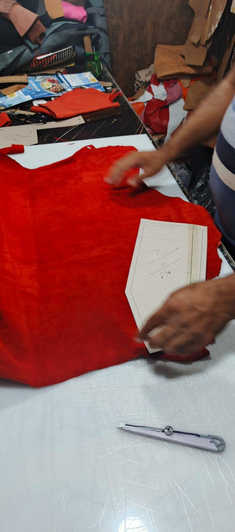 Puede incluir: Una pieza grande de cuero rojo vibrante extendida sobre una superficie blanca, con una plantilla de patr&oacute;n de papel beige encima. La plantilla presenta marcas escritas a mano y un borde superior puntiagudo. Un c&uacute;ter plateado descansa cerca.