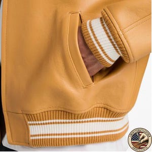 May include: Close-up of a mustard yellow leather jacket with a ribbed knit cuff and hem. The cuff and hem feature white stripes. A hand is in the jacket pocket. The jacket has a pocket on the front. The logo "Liberty Craft Leather" is visible.