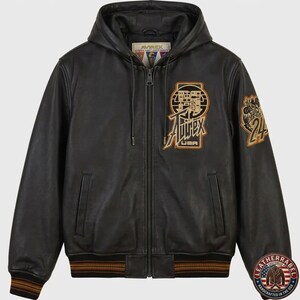 May include: A black leather hooded jacket with a full-length front zipper, ribbed cuffs and hem with brown stripes, and two side pockets. The chest features an embroidered Avirex USA logo patch, and the left sleeve has a circular 24 patch.