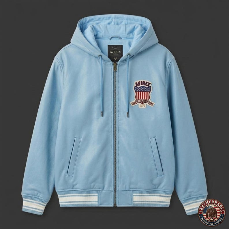 Pu&ograve; includere: Giacca in pelle azzurra con cappuccio e chiusura a zip. La giacca presenta una toppa con la scritta "AVIREX" e un disegno della bandiera americana. I polsini e l'orlo hanno dettagli a righe bianche e blu. Due tasche frontali.