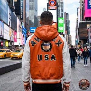 K&ouml;nnte beinhalten: Orange-wei&szlig;e Lederjacke mit Kapuze, mit den Schriftz&uuml;gen "AVEX" und "USA" sowie einem "All Americans 1975" Patch. Die Jacke hat wei&szlig;e &Auml;rmel und gestreifte B&uuml;ndchen. Das Bild ist in einer Stadt mit gelben Taxis und Werbetafeln aufgenommen.