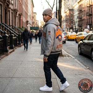 Puede incluir: Una chaqueta universitaria gris claro con capucha, bordado naranja y azul de los New York Knicks en la espalda, gorro de punto marr&oacute;n, vaqueros azul oscuro y zapatillas blancas. La chaqueta tiene pu&ntilde;os de canal&eacute; a rayas y un parche peque&ntilde;o.