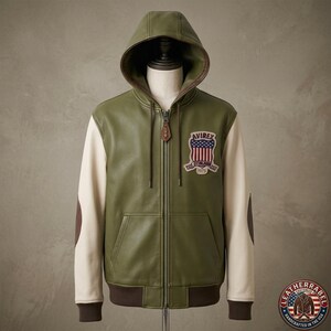 May include: An olive green leather hooded jacket with cream-colored sleeves and brown elbow patches. The front features a full-length zipper with a leather pull tab and an embroidered patch on the chest displaying the text AVIREX and 1975.