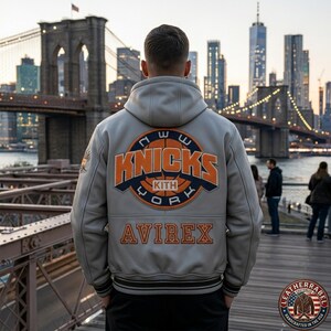 Puede incluir: Una chaqueta universitaria con capucha de color gris claro que presenta un gran parche circular en la espalda con el texto en naranja y azul marino "NEW YORK KNICKS KITH" y un gr&aacute;fico de baloncesto. "AVIREX" est&aacute; bordado en naranja.