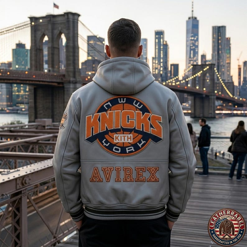 Puede incluir: Una chaqueta universitaria con capucha de color gris claro que presenta un gran parche circular en la espalda con el texto en naranja y azul marino "NEW YORK KNICKS KITH" y un gr&aacute;fico de baloncesto. "AVIREX" est&aacute; bordado en naranja.