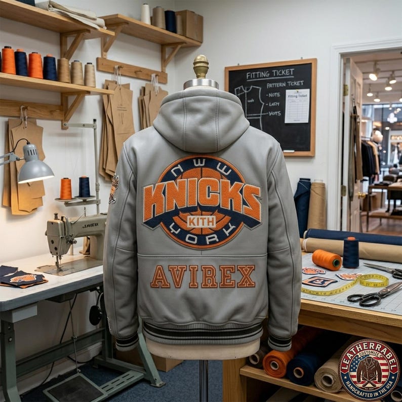 Puede incluir: Una chaqueta universitaria con capucha de cuero gris que presenta un gran logotipo circular naranja y azul marino de Knicks Kith New York en la espalda. La palabra AVIREX est&aacute; bordada en naranja debajo del logo. Tiene pu&ntilde;os y dobladillo acanalados.