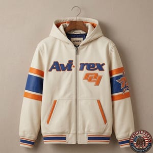 Pode incluir: Casaco com capuz com zíper creme com o nome da marca "Avi-rex" em letras azuis e laranja. O casaco apresenta listras laranja e azul nas mangas e um gráfico de estrela. O casaco tem bainha e punhos canelados.