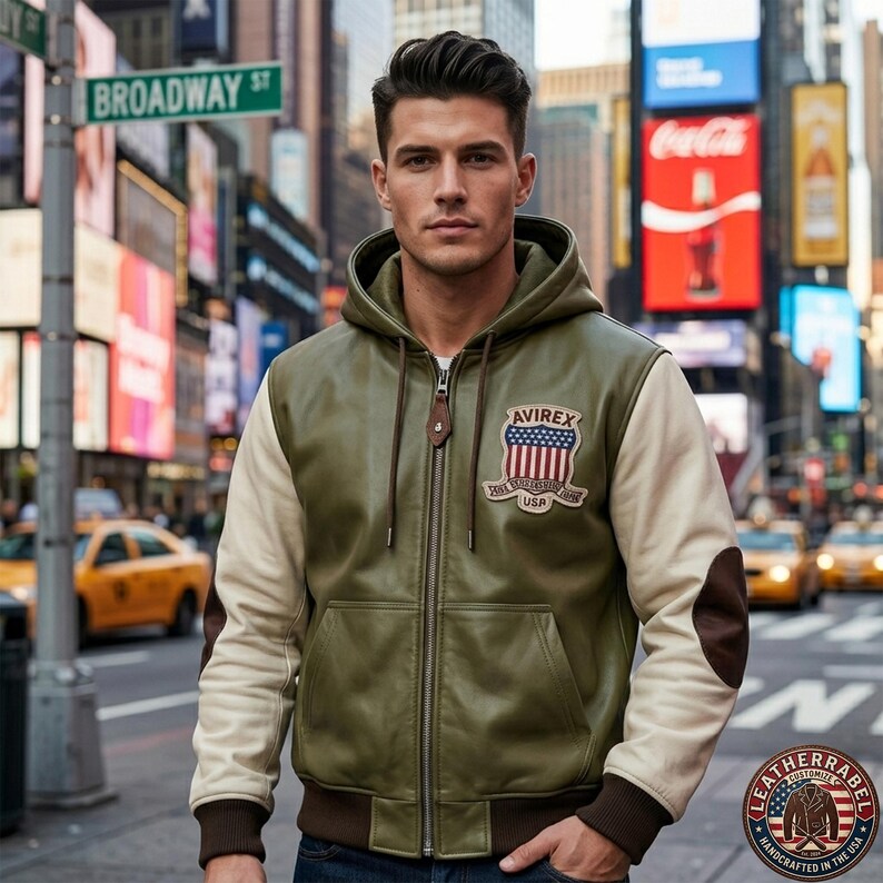 May include: An olive green leather bomber jacket with a hood, featuring cream-colored sleeves, dark brown elbow patches, and a ribbed hem. A patch on the chest displays the text AVIREX above a shield with stars and stripes and the letters USA.