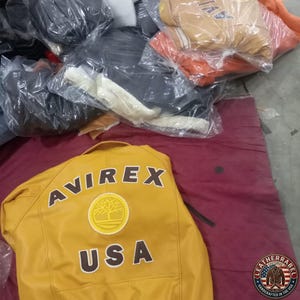 May include: Mustard yellow leather jacket with the word "AVIREX" and "USA" printed on the back. The jacket has a circular emblem. Other jackets are visible in the background, wrapped in plastic.