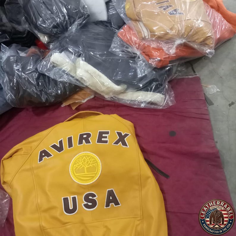 May include: Mustard yellow leather jacket with the word "AVIREX" and "USA" printed on the back. The jacket has a circular emblem. Other jackets are visible in the background, wrapped in plastic.