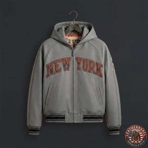 Puede incluir: Una chaqueta gris con capucha y cremallera frontal completa, que presenta el texto NEW YORK en letras arqueadas con bordes oscuros en el pecho. La prenda incluye pu&ntilde;os y un dobladillo acanalados con rayas negras y forro interior.