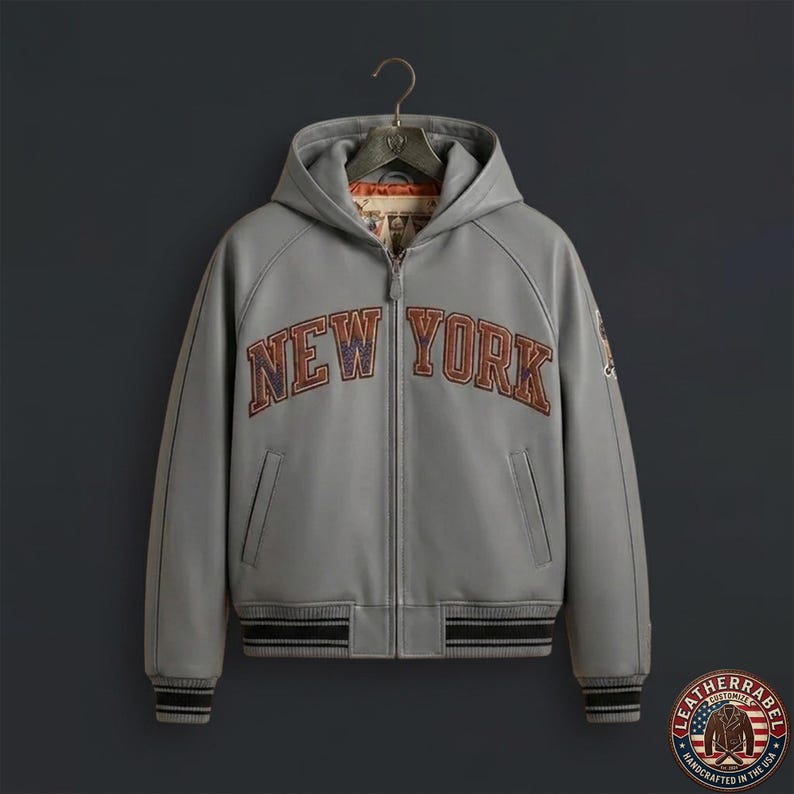 Puede incluir: Una chaqueta gris con capucha y cremallera frontal completa, que presenta el texto NEW YORK en letras arqueadas con bordes oscuros en el pecho. La prenda incluye pu&ntilde;os y un dobladillo acanalados con rayas negras y forro interior.