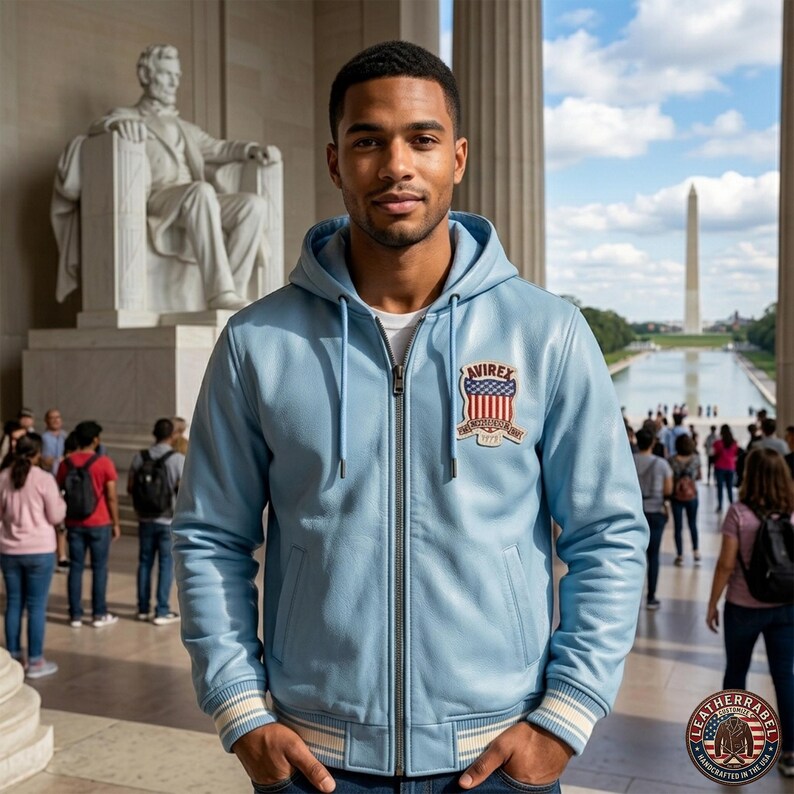 Pu&ograve; includere: Giacca in pelle azzurra con cappuccio e chiusura a zip. La giacca presenta una toppa con la scritta "AVIREX" e un design patriottico. La giacca ha polsini e orlo a righe bianche e blu. Lo sfondo mostra il Lincoln Memorial e il Monumento a Washington.