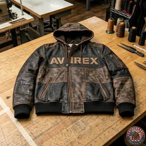 May include: A brown and black leather jacket with a hood and the word "AVIREX" across the chest. The jacket has a zip closure, two front pockets, and black ribbed cuffs and hem. The jacket is on a wooden surface.