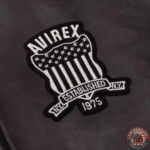 May include: A black and white embroidered patch with the text "AVIREX" at the top, and "ESTABLISHED NY 1975" on a banner. The patch has a shield design with vertical stripes and stars, and is likely for a jacket or apparel.