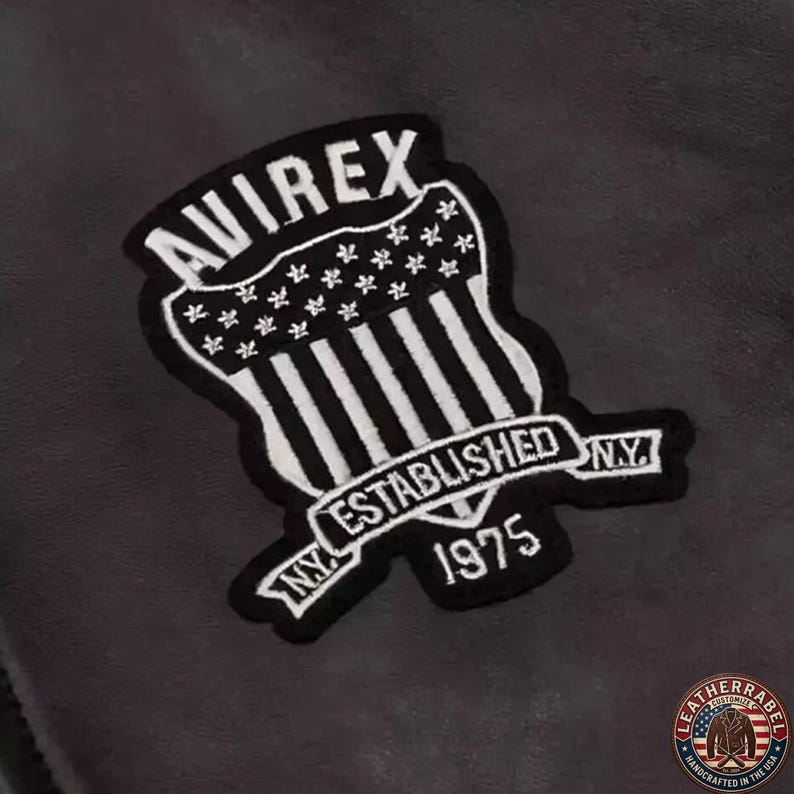 May include: A black and white embroidered patch with the text "AVIREX" at the top, and "ESTABLISHED NY 1975" on a banner. The patch has a shield design with vertical stripes and stars, and is likely for a jacket or apparel.