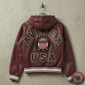 Puede incluir: Una chaqueta de cuero burdeos con capucha que presenta el texto AVIREX USA en letras negras grandes en la parte posterior. Un parche circular en el centro muestra un gr&aacute;fico de la bandera estadounidense con el texto ALL AMERICANA ESTABLISHED 1975. La chaqueta tiene pu&ntilde;os y dobladillo acanalados con rayas blancas y burdeos.